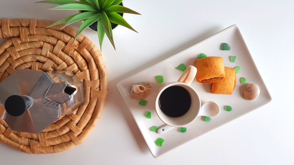 vassoio di ceramica decorato con vetrini e conchiglie, tazzina di caffè, cucchiaino di ceramica e biscotti, con una caffettiera a sinistra.